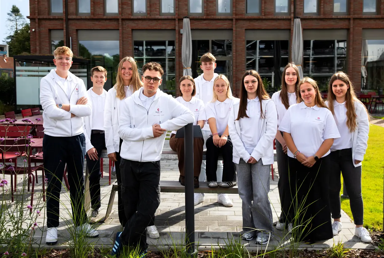Eine Gruppe junger Menschen steht in einem Gartenbereich vor einem Gebäude. Sie tragen weiße Poloshirts und lächeln in die Kamera. Einige sitzen auf einer Bank, andere stehen daneben. Im Hintergrund sind Pflanzen und eine Terrasse sichtbar.
