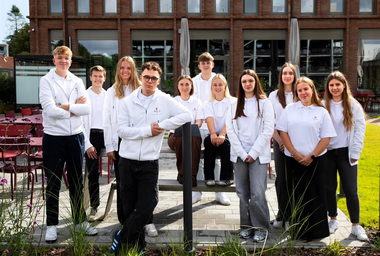 Eine Gruppe junger Menschen steht in einem Gartenbereich vor einem Gebäude. Sie tragen weiße Poloshirts und lächeln in die Kamera. Einige sitzen auf einer Bank, andere stehen daneben. Im Hintergrund sind Pflanzen und eine Terrasse sichtbar.