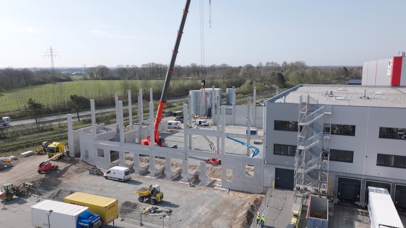 Construction workers are assembling a new building, using cranes and machinery to position concrete columns and beams. Several vehicles are present on the site, and the surrounding landscape includes fields and trees under a clear sky.