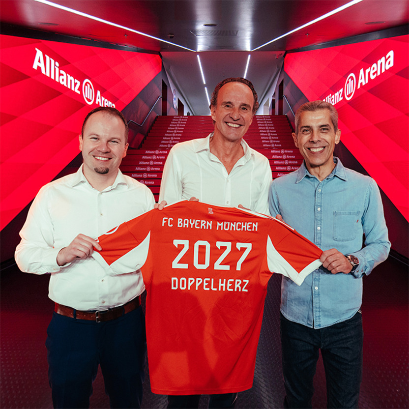Drei Personen stehen in der Allianz Arena und halten ein rotes Trikot des FC Bayern München. Auf dem Trikot steht „2027 DOPPELHERZ“. Der Hintergrund ist in rötlichen Tönen gehalten, die Arena-Beschilderung ist deutlich sichtbar.