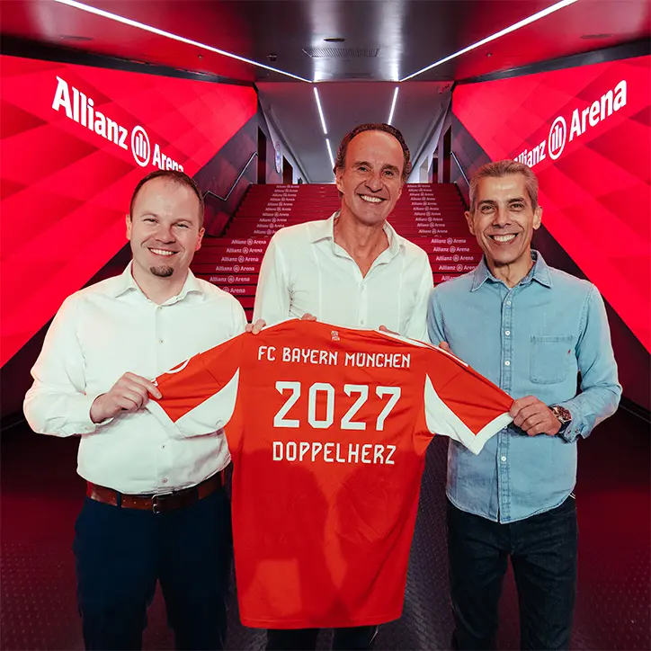 Drei Personen stehen in der Allianz Arena und halten ein rotes Trikot des FC Bayern München. Auf dem Trikot steht „2027 DOPPELHERZ“. Der Hintergrund ist in rötlichen Tönen gehalten, die Arena-Beschilderung ist deutlich sichtbar.
