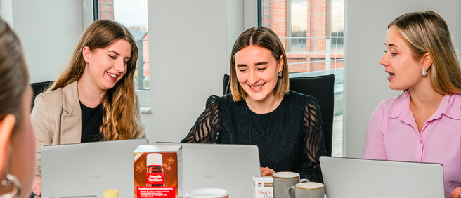 Drei Frauen sitzen an einem Tisch und arbeiten an Laptops. Sie lächeln und unterhalten sich, während auf dem Tisch einige Getränke und Snacks stehen. Die Atmosphäre wirkt freundlich und entspannt.