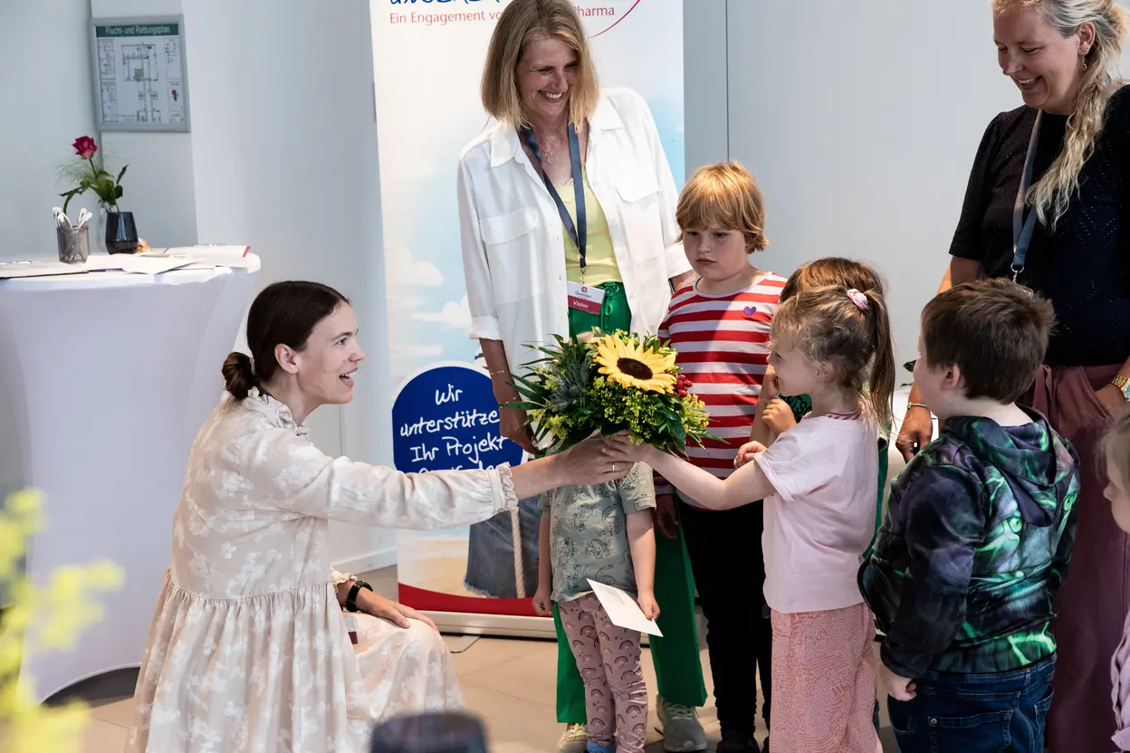 A woman kneels to receive a bouquet of flowers from a young girl, surrounded by several other children. An adult smiles in the background, with a banner visible that suggests support for community projects. The atmosphere appears warm and engaging.