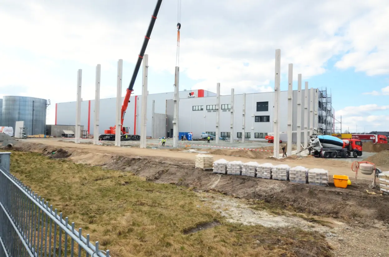 Baustelle des Queisser-Logistikzentrums mit Kran, Betonstützen im Aufbau, LKWs und Baumaterialien im Vordergrund.