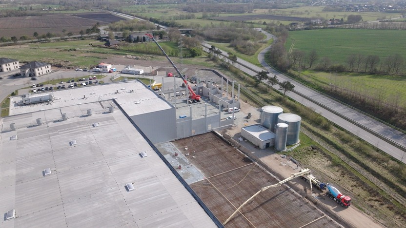 An aerial view of a construction site featuring a large building with steel support columns being erected. Nearby, construction vehicles are present, and silos are visible. The surrounding landscape includes fields and a road. The scene indicates ongoing industrial development.