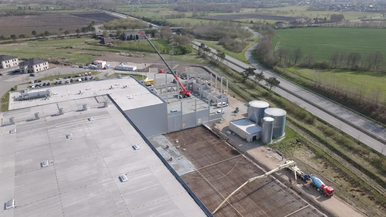 An aerial view of a construction site featuring a large building with steel support columns being erected. Nearby, construction vehicles are present, and silos are visible. The surrounding landscape includes fields and a road. The scene indicates ongoing industrial development.